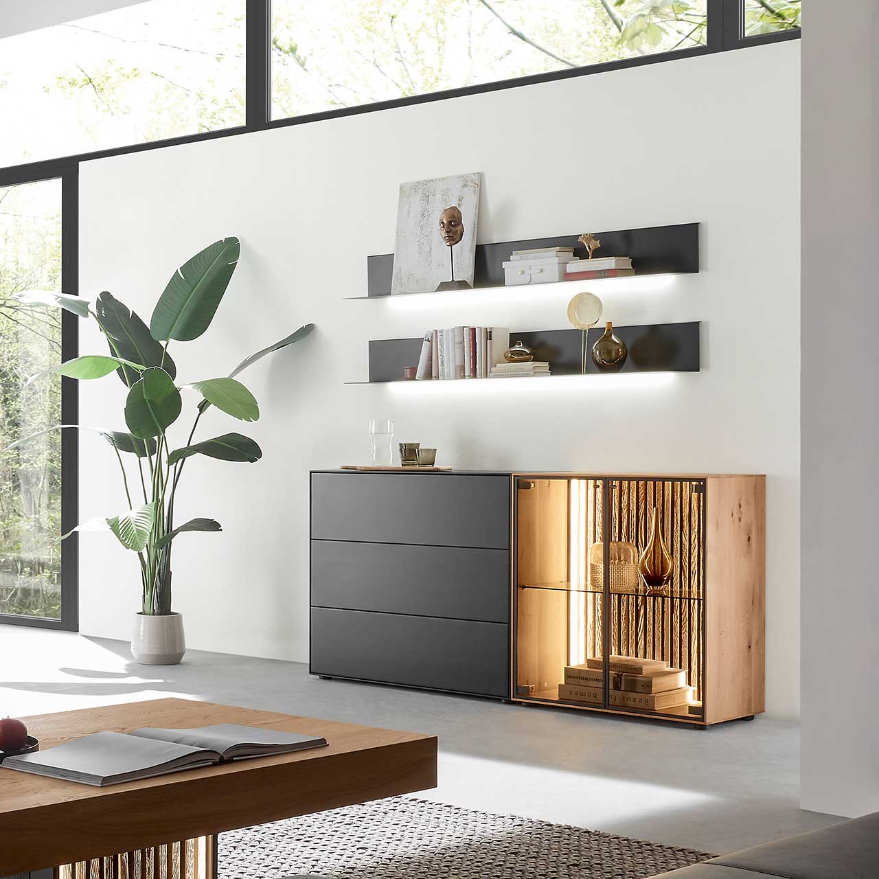 Modernes Wohnzimmer mit beleuchtetem Sideboard aus Birke anthrazit mit Glastüren, Lamellenrückwand und dunklen Schubkästen, darüber hängend zwei Metallregale in anthrazit. Im Vordergrund der passende Holz-Couchtisch.