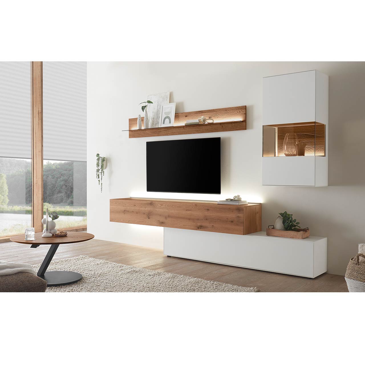 Wohnzimmer mit moderner Wohnwand aus weiß lackierter Birke und dunkler Kerneiche bestehend aus langem, rechteckigen Hängelowboard, weißemUnterschrank, weißer Hängevitrine und Wandregal mit Glasablageboden. Davor ein runder Couchtisch.