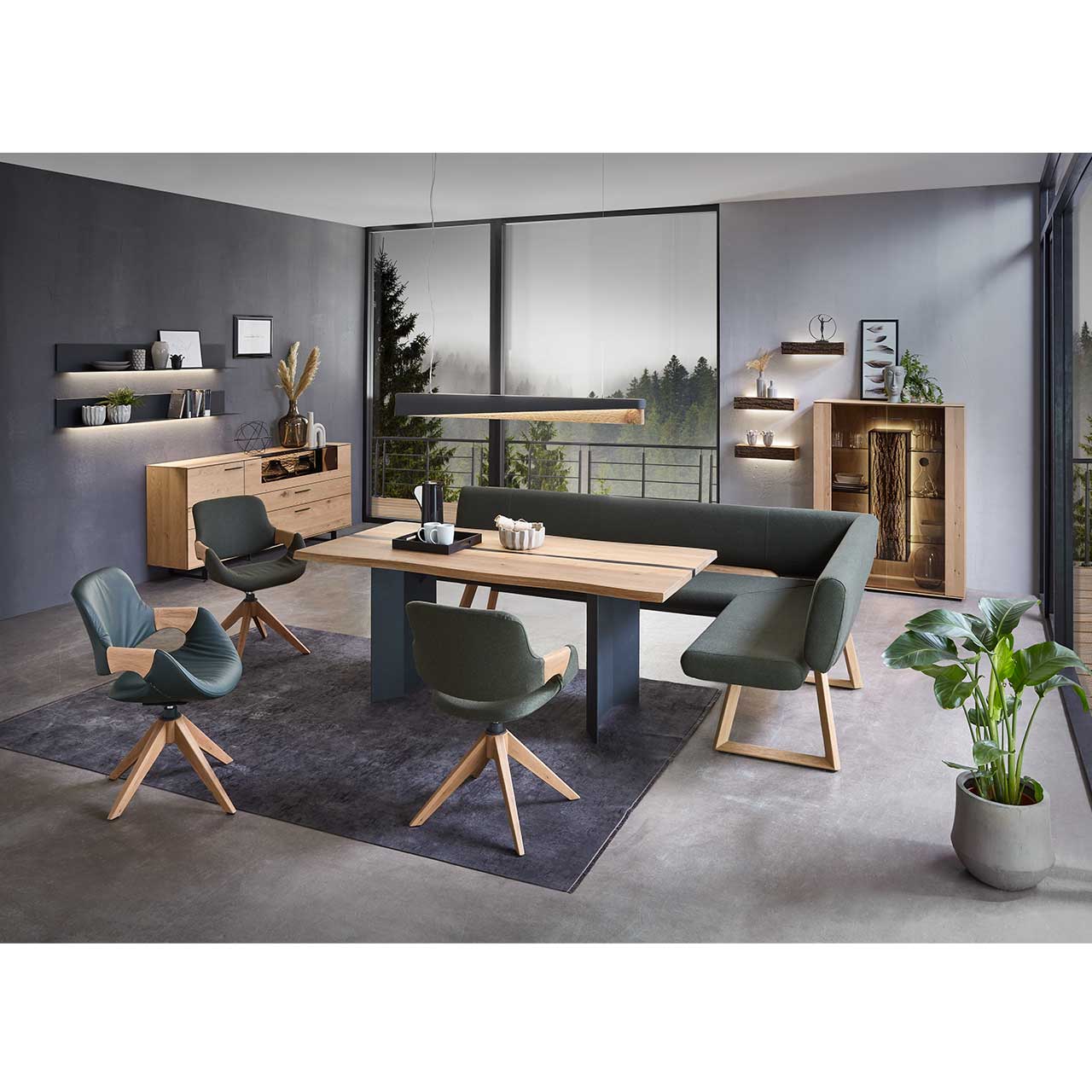 Modernes Esszimmer mit massivem, rechteckigen Holztisch, umgeben von drei Stühlen mit dunkelgrünem Lederbezug auf Holzgestell und der passenden Ecksitzbank. Hinten an der Wand stehend die passenden Esszimmermöbel.