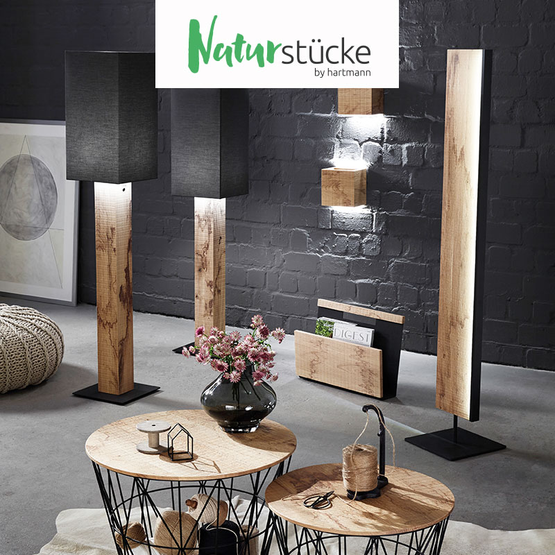 Hartmann NATURSTÜCKE - extravagante Wohnaccessoires aus massiver Birke, Esche oder Kerneiche kombiniert mit Glas und Metall. Ambientebild zeigt verschiedene Stehlampen, Zeitungsständer, Wandleuchten, Beistelltische aus hochwertigem Holz.
