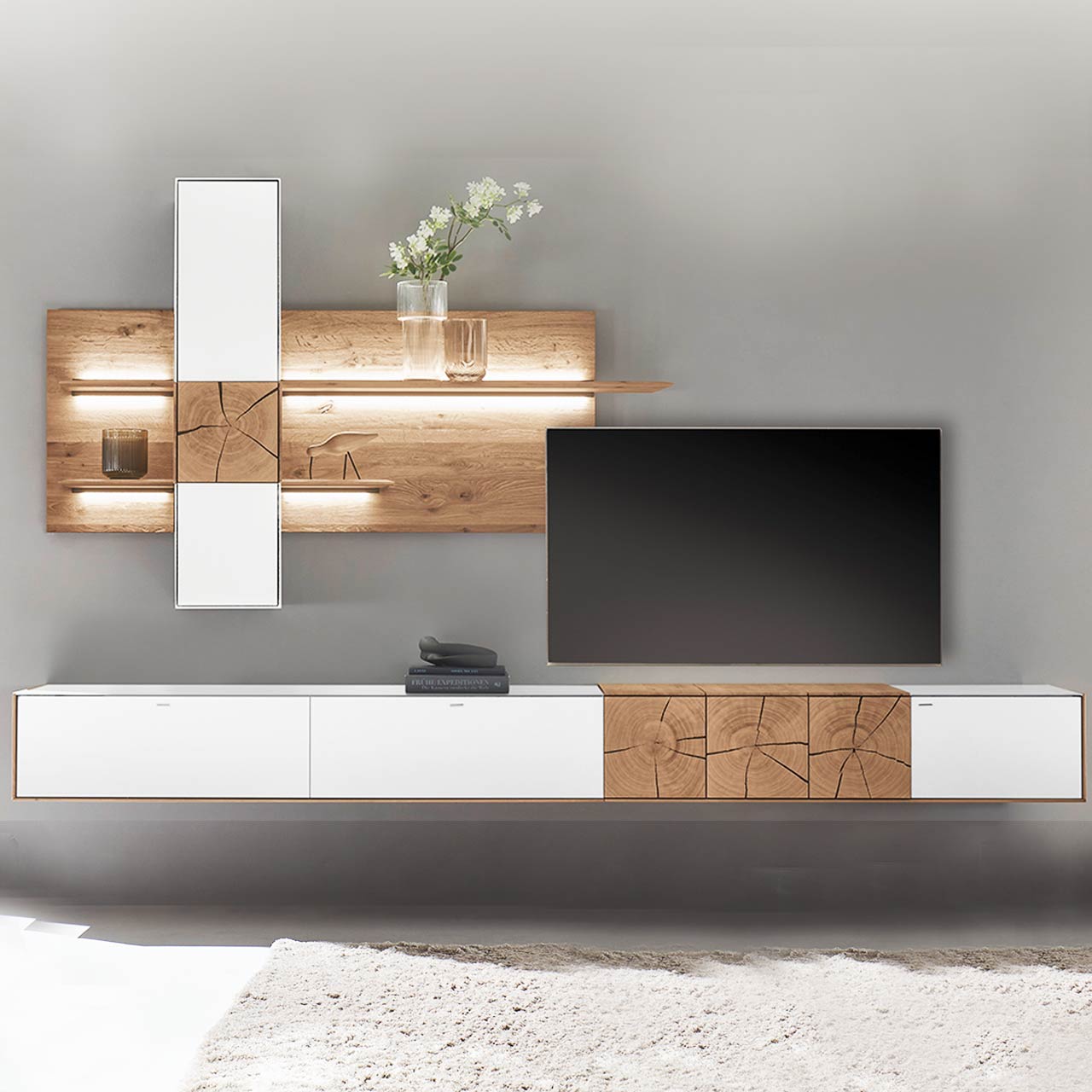 Hartmann CAYA - Wohnwand 7170 Nr. V72W - dreiteilig Moderne Wohnwand aus massiver Kerneiche mit Mattglas weißen Fronten. Unten ein langes Hängelowboard mit dekorativer Holzapplikation, oben eine Wandpaneel-Kombination mit Regalböden, Schrank und indirekter Beleuchtung, dazwischen hängender Fernseher.