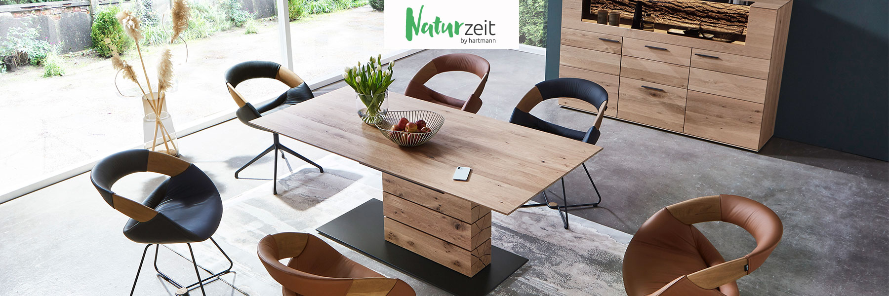Hartmann NATURZEIT - moderne Esszimmermöbel aus massivem Holz kombiniert mit Glas und Metall. Ambientebild zeigt rechteckigen Esstisch mit massiver Holzsäule umgeben von sechs komfortablen Stühlen in Leder mit Drahtgestell in Metall anthrazit.