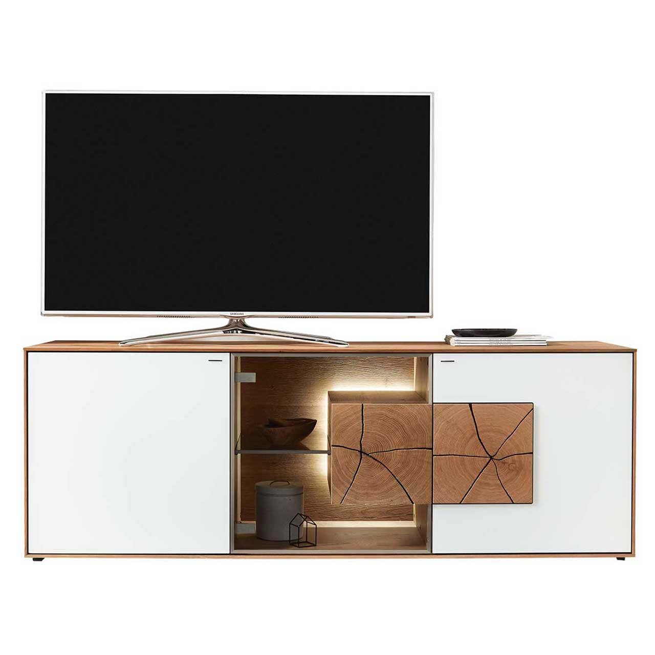 Hartmann CAYA - Lowboard 7170-3175W Großer TV platziert auf modernem Lowboard aus massiver Kerneiche Umato mit Fronten aus Mattglas weiß, mittig Glastür mit Glasboden und beleuchtetem Hirnholzblock.
