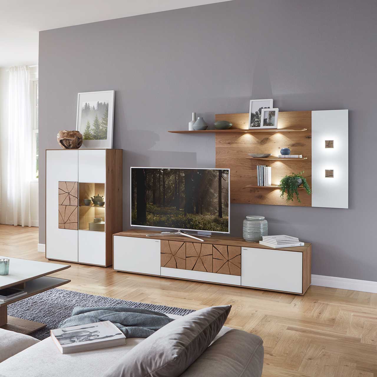 Wohnzimmer mit moderner dreiteiliger Massivholz Wohnwand mit Mattglas weißen Fronten und dekorativen Hirnholzscheiben: Langes Lowboard mit Hirnholz und Kufen, Wandpaneel mit Regalböden, Highboard mit beleuchtetem Glasausschnitt und Hirnholz.