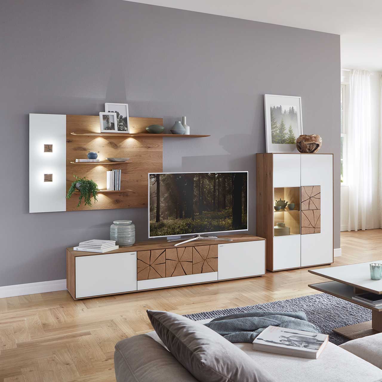 Wohnzimmer mit moderner dreiteiliger Massivholz Wohnwand mit Mattglas weiß Fronten und dekorativen Hirnholzscheiben: Langes Lowboard mit Hirnholz und Kufen, Wandpaneel mit Regalböden, Highboard mit beleuchtetem Glasausschnitt und Hirnholz.