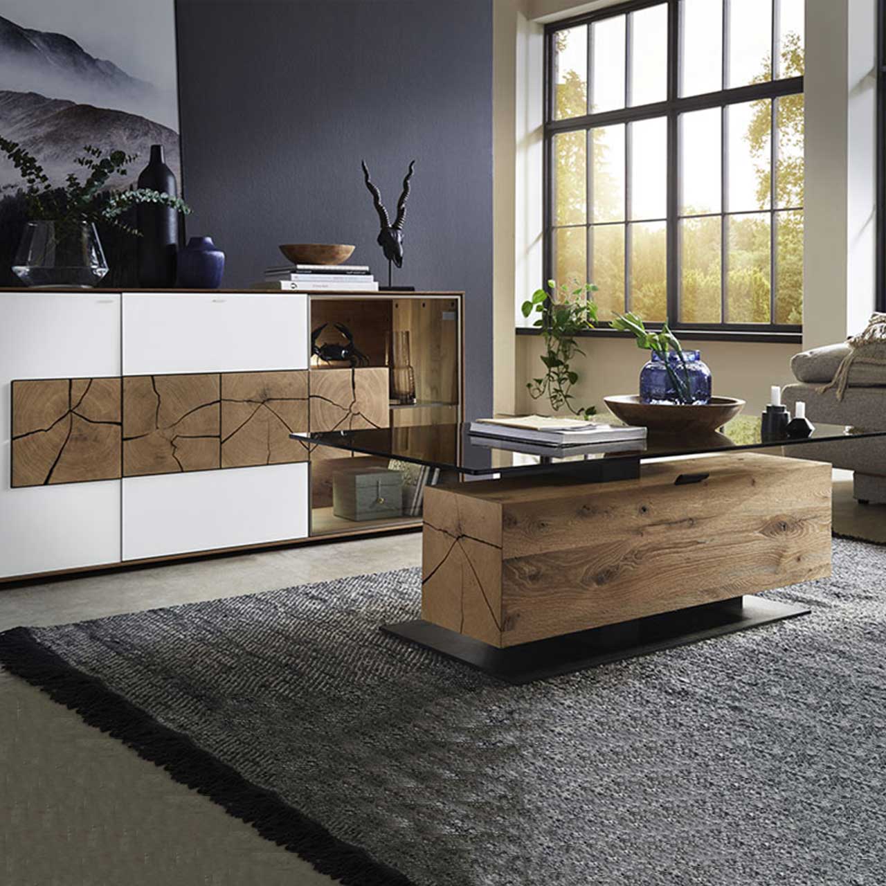Bildausschnitt eines Wohnzimmers mit modernem Lowboard aus massiver Kerneiche Umato mit weißfarbenen Mattglasfronten, drei rechteckigen Hirnholzscheiben und Glastür mit beleuchtetem Glasboden und Hirnholzblock, davor passender Couchtisch.