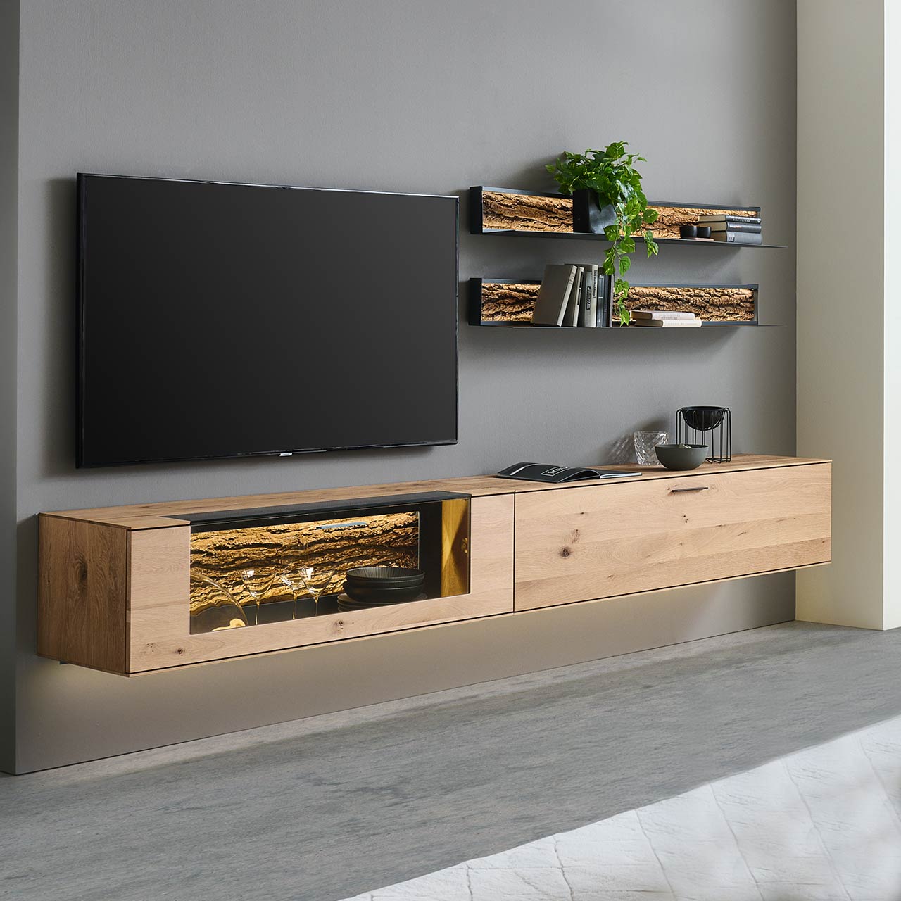 Moderne vierteilige Wohnwand bestehend aus zwei Hängelowboards aus massiver Kerneiche Natur, eines mit beleuchtetem Glasausschnitt und Rinden-Akzent, dazu zwei moderne Metall-Wandregale mit sichtbarer Baumrinde.