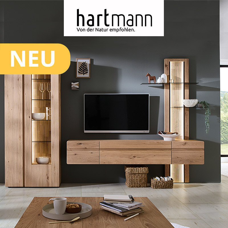 Hartmann VIDA - Massivholzmöbel aus Kerneiche Natur kombiniert mit geschoppten Rückwänden und Holzeinkerbungen. Ambientebild zeigt Hängelowboard, Wandpaneel mit beleuchteten Glasböden und eine Standvitrine - vor dunkelgrauer Wand.