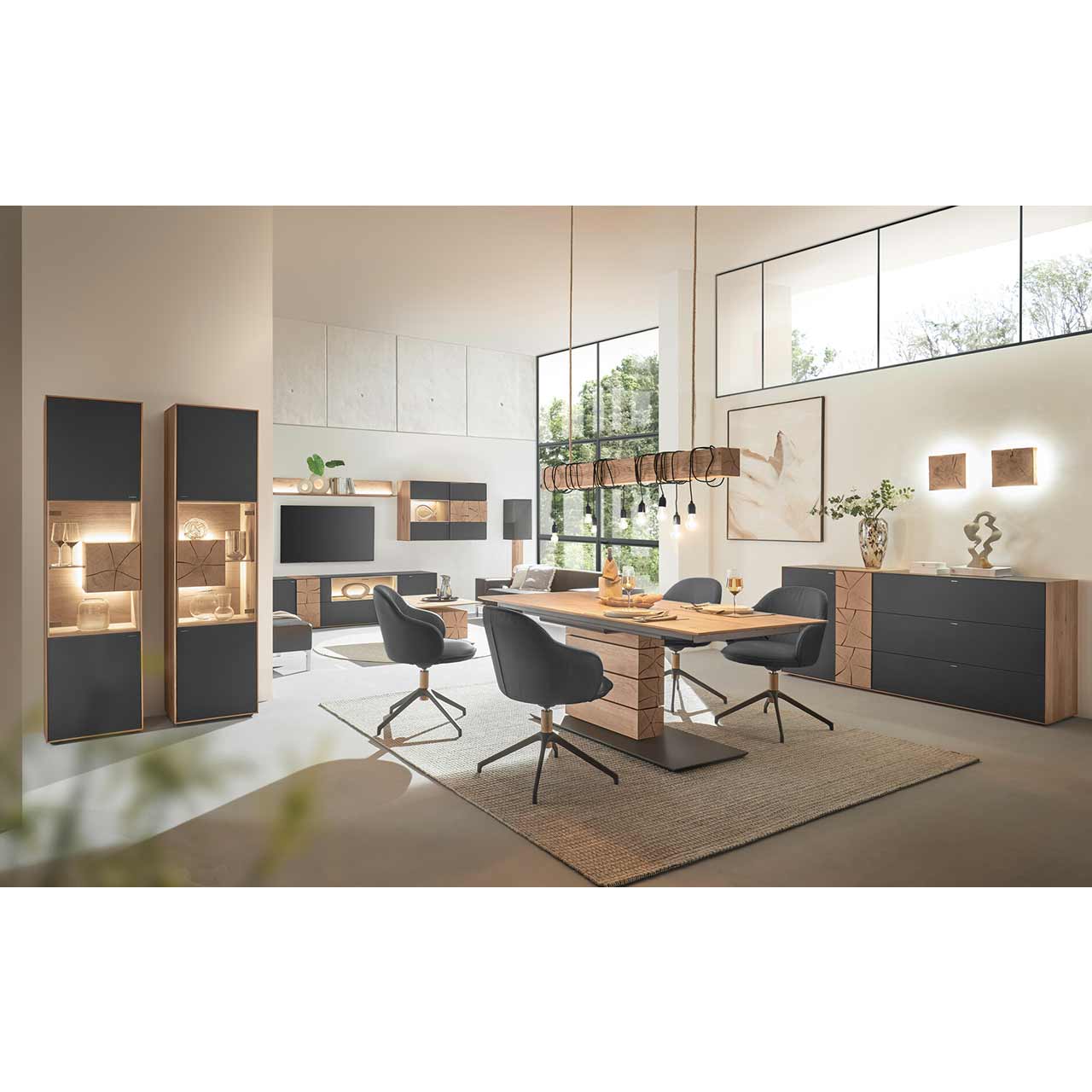 Modern eingerichtetes Esszimmer mit Esstisch aus massivem Holz umgeben von vier Stühlen mit schwarzem Lederbezug und Metallgestell. Darüber passende Balkenleuchte. Links ein Sideboard mit Mattglas Anthrazit Front, rechts zwei passende Standvitrinen.