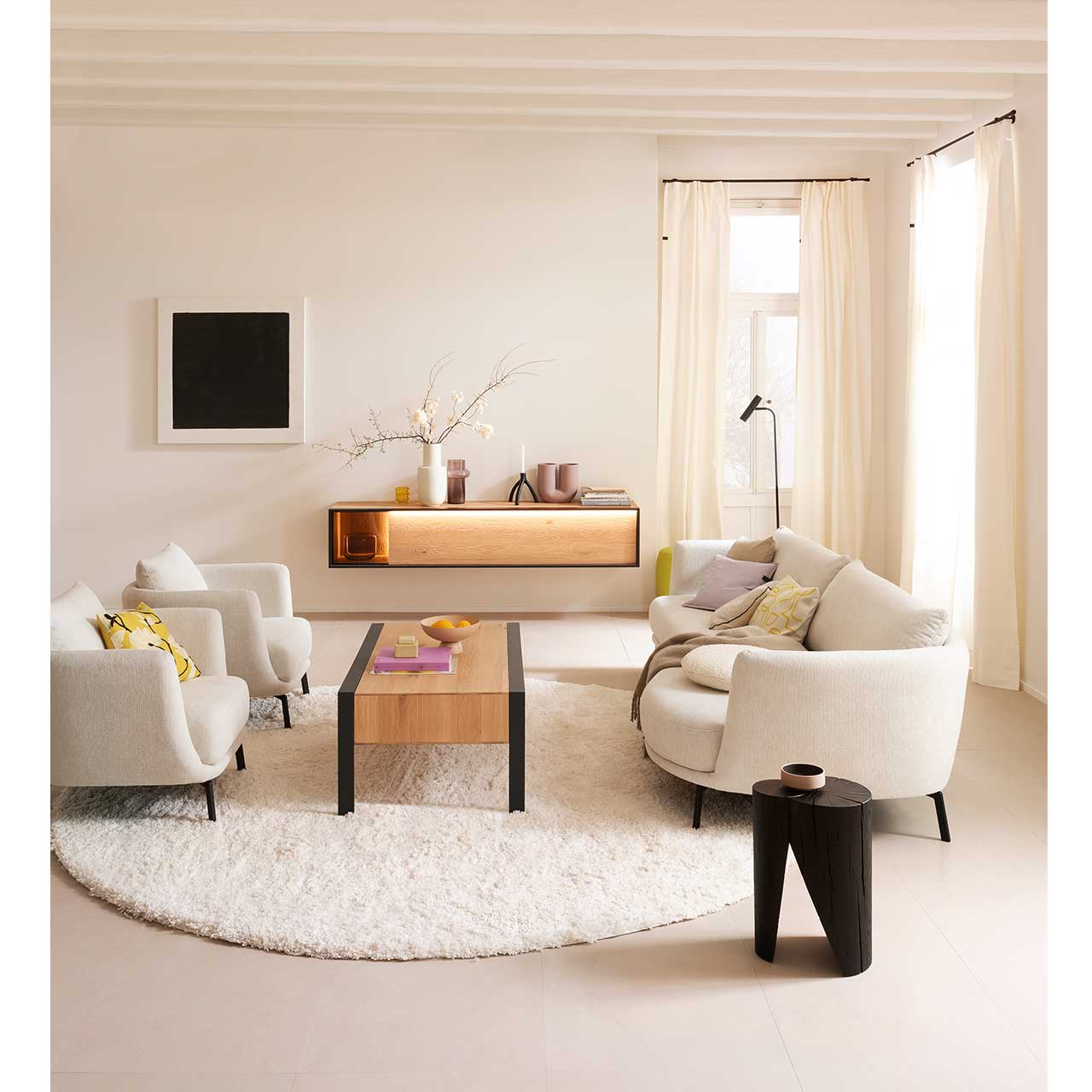 Helles Wohnzimmer mit beleuchtetem Hängelowboard aus massiver sandgestrahlter Kerneiche Natur eingefasst in Metallrahmen anthrazit und beleuchtetem Glasausschnitt an hell-beiger Wand. Davor steht ein rechteckiger Holz-Couchtisch auf Metallfüßen anthrazit.
