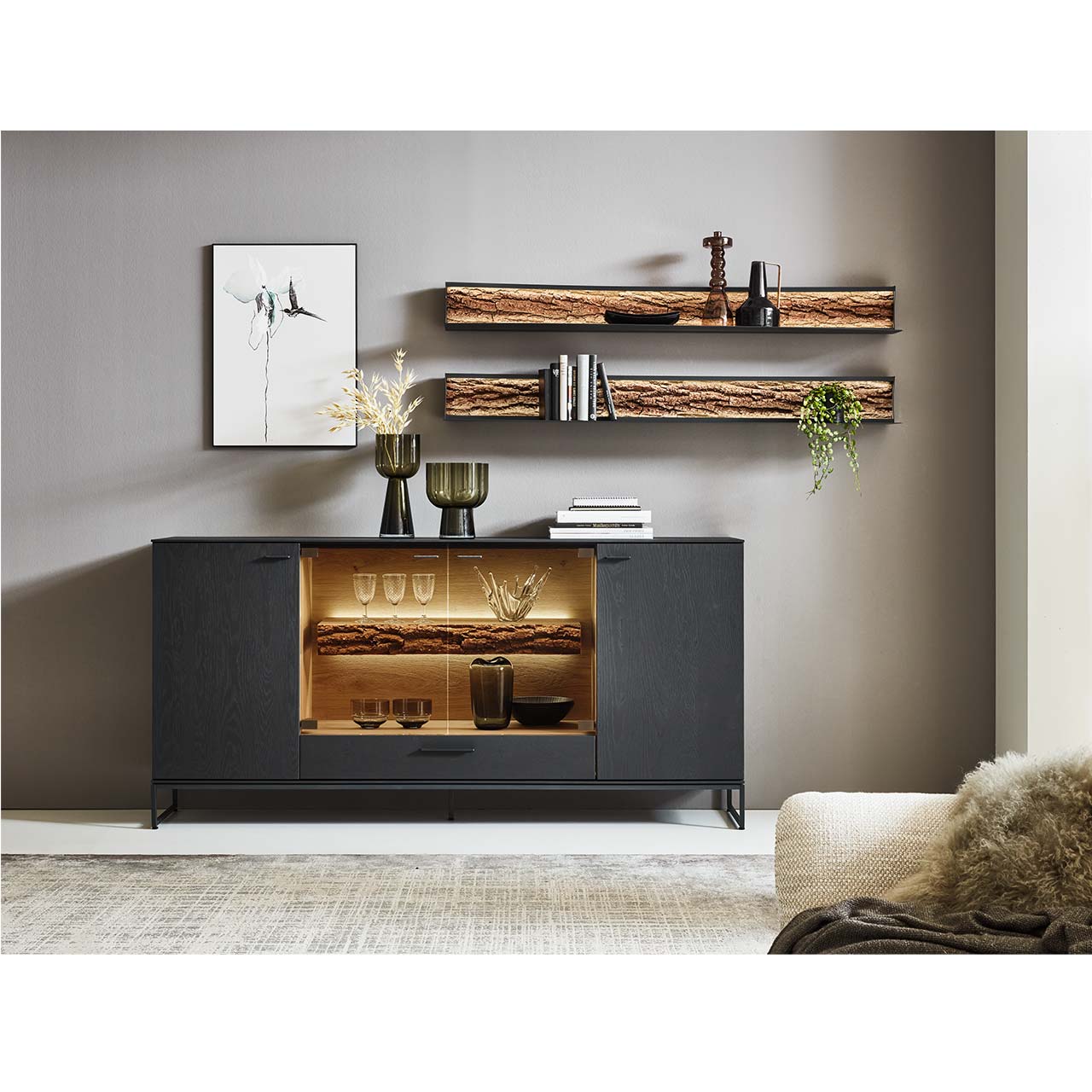 Wohnzimmer mit modernem Sideboard aus Kerneiche Natur in anthrazit gebeizt, mit beleuchtetem Glasausschnitt und Rinden-Akzent- auf Metallkufen. Darüber zwei hängende Metallregale mit Rinden-Akzent.