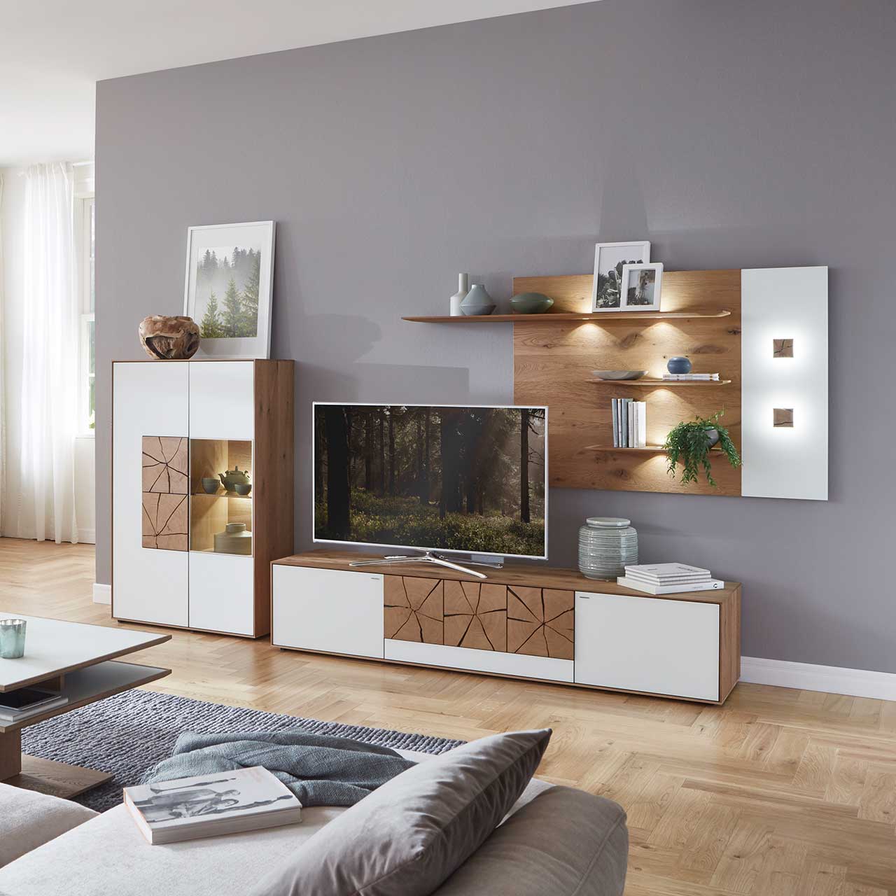 Milieubild Hartmann CAYA - Standelement 7170-7097W li. Wohnzimmer mit moderner Massivholz Wohnwand mit Mattglas weiß Fronten und dekorativen Hirnholzscheiben: Langes Lowboard mit Hirnholz, Wandpaneel mit zwei Regalböden und Highboard mit beleuchtetem Glasausschnitt und Hirnholz.