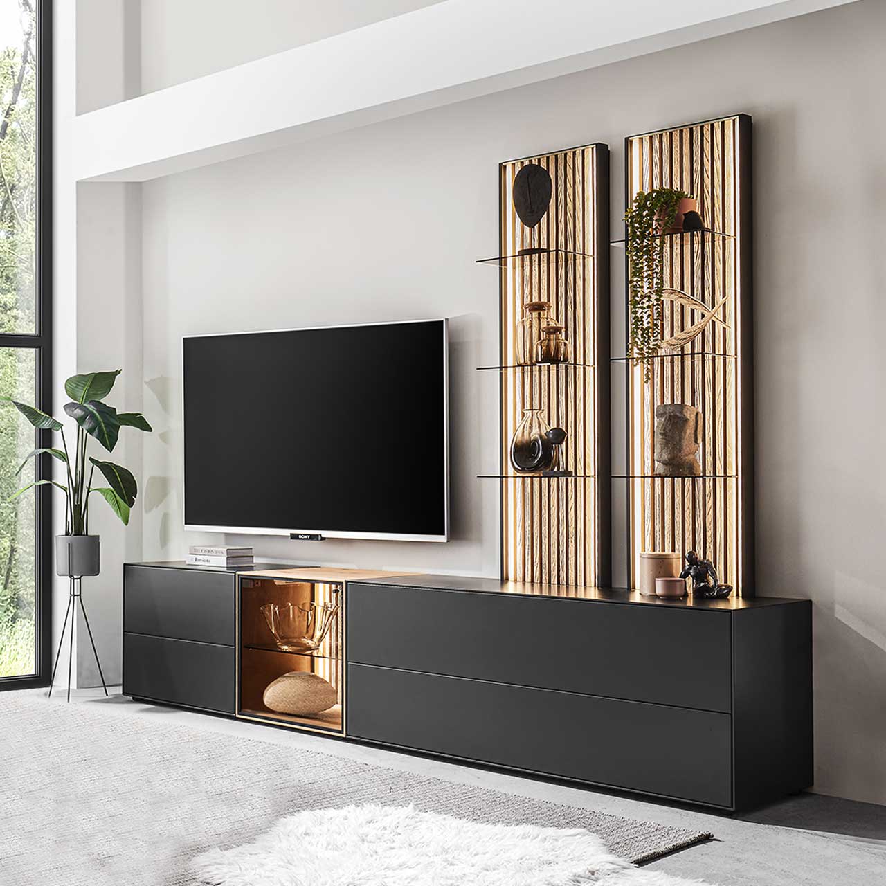 Wohnzimmer mit Fernseher und moderner fünfteiliger Wohnwand mit Lowboards in Birke anthrazit und Glastüren sowie zwei hohen Wandpaneelen mit hellen Holzlamellen.