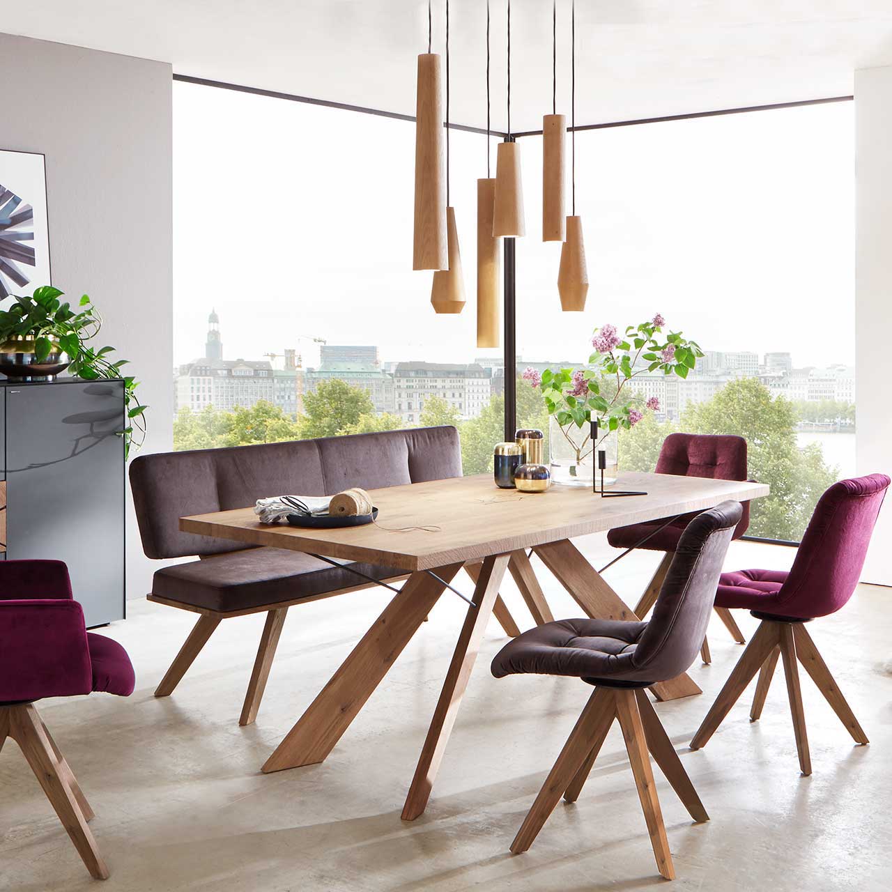 Modernes Esszimmer mit Tisch aus massivem Holz mit sternförmigem Holzgestell, vier Stühlen und Sitzbank in bordeaux-braunem Bezug mit Holzgestell, darüber hängend Pendelleuchte mit sechs unterschiedliche geometrische Holzkörpern.