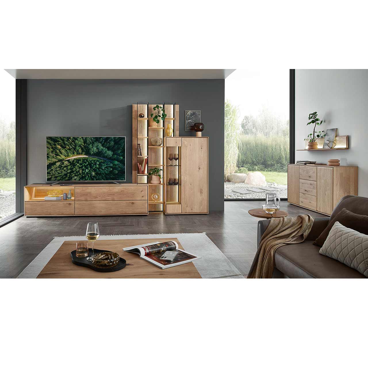 Modernes Wohnzimmer mit Massivholzwohnwand bestehend aus einem Highboard mit Glastür, einem Hochregal mit Holzböden, beleuchteten Glasseiten und geschroppter Rückwand und einem Lowboard mit Nische. Davor eckiger Couchtisch, rechts passendes Sideboard.