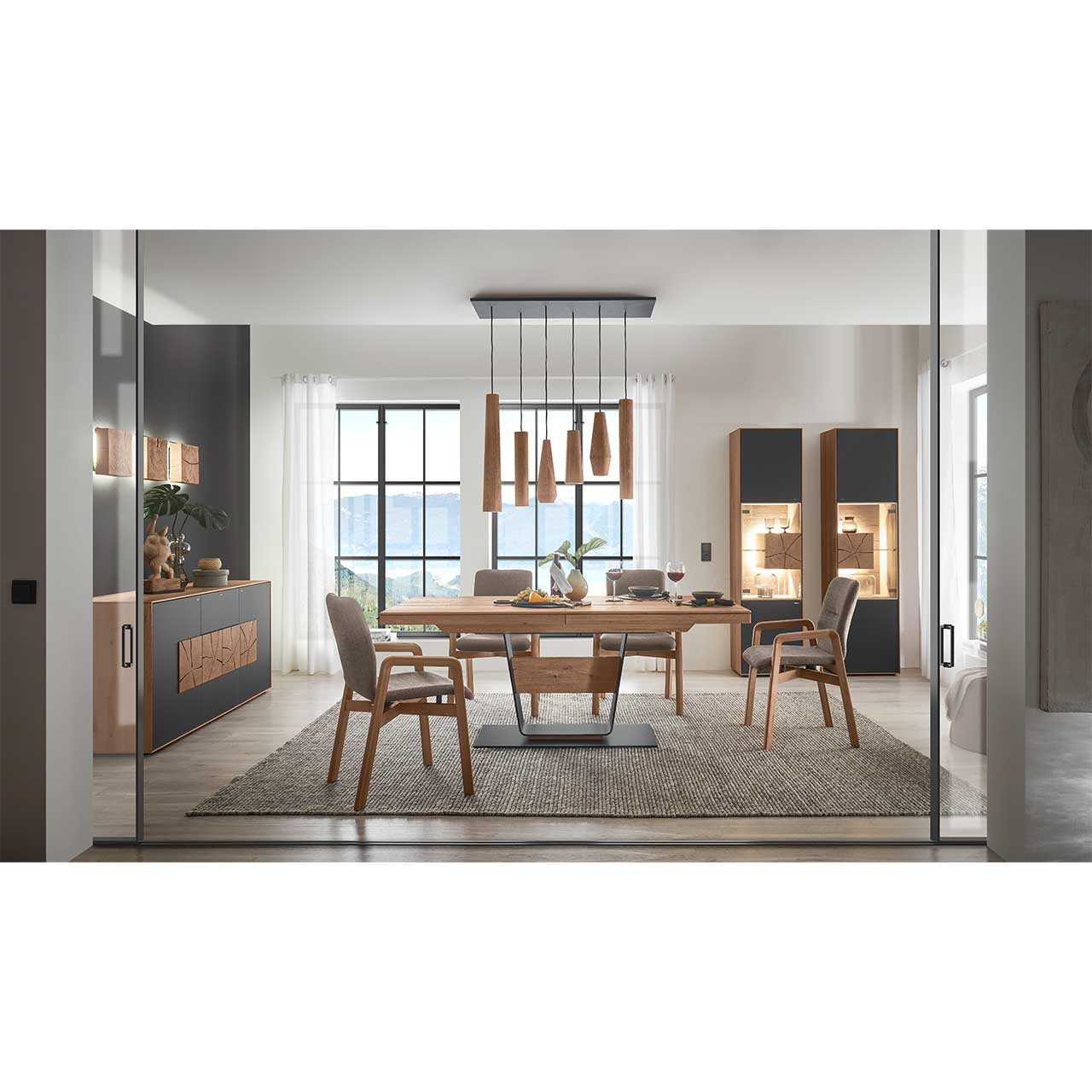 Modernes Esszimmer mit rechteckigem Massivholz Tisch und vier Stühlen mit Holzgestell. Über dem Tisch hängt eine Pendelleuchte mit Holz-Lampenschirmen. Im Hintergrund links steht ein Sideboard in Anthrazit, rechts zwei passende Standvitrinen.