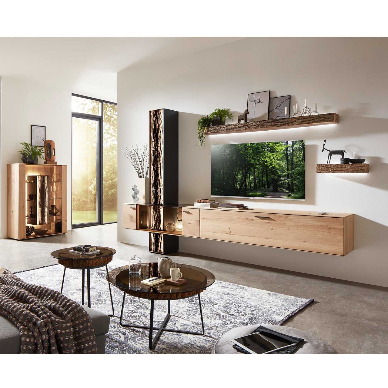 Wohnzimmer mit moderner Massivholz Wohnwand. Links das passende Highboard mit Glasfront. Im Vordergrund ein kleiner und ein großer Beistelltisch mit runder Glasplatte auf echter Baumscheibe und filigranem Metallgestell in Anthrazit.