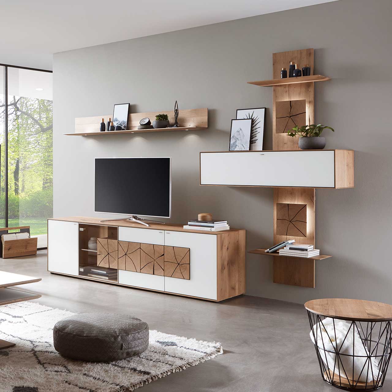 Milieubild Hartmann CAYA - Hängeelement 7170-1135W Wohnzimmer mit TV und moderner Wohnwand aus massiver Kerneiche Umato mit Mattglas weiß Fronten: Lowboard mit Glastür und Hirnholzscheiben, länglicher Hängeschrank eingefasst in beleuchtetes Wandpaneel mit Ablageböden, daneben Hängeregal aus Holz.