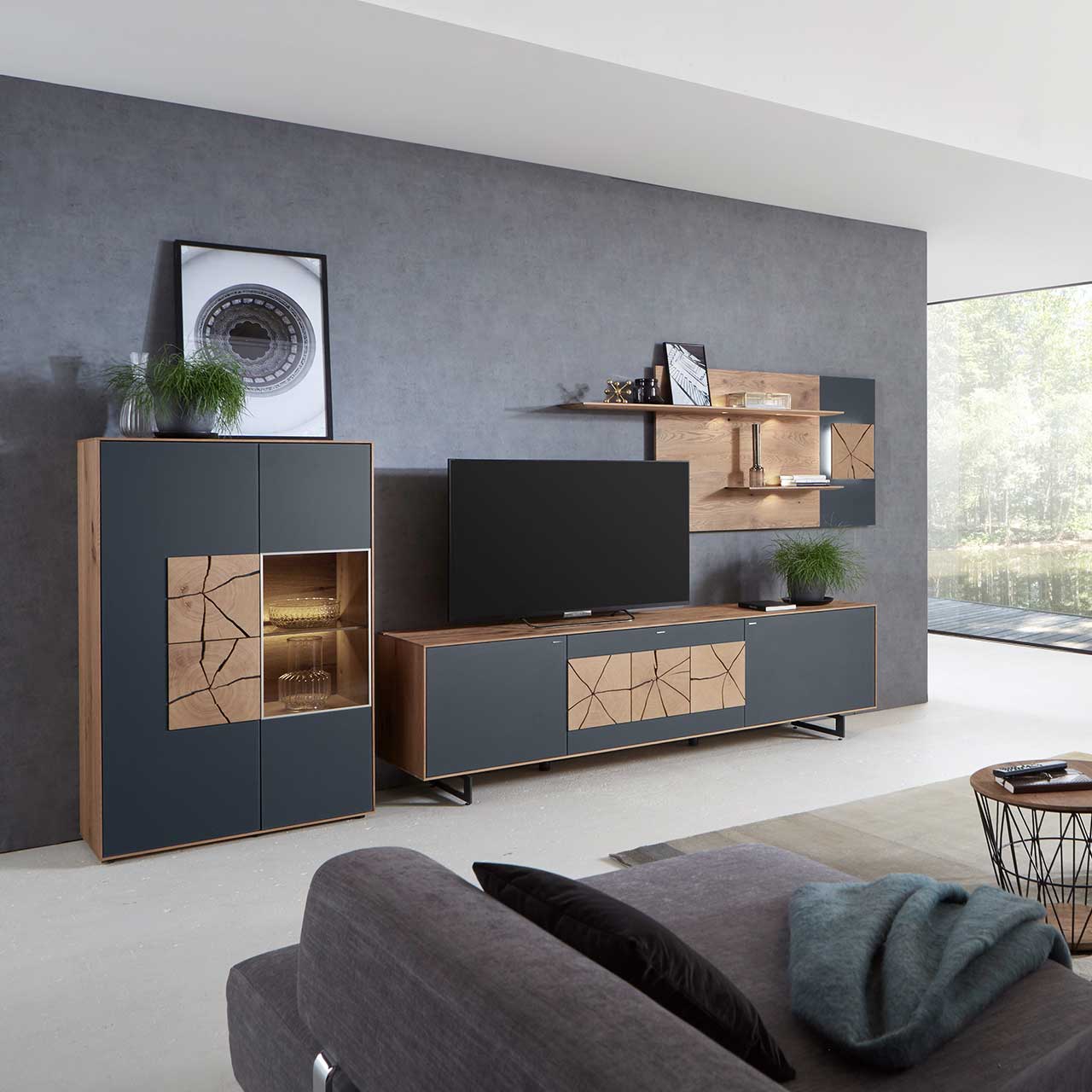 Milieubild Hartmann CAYA - Hängeelement 7170-7091A li. Wohnzimmer mit moderner dreiteiliger Massivholz Wohnwand mit Mattglas Anthrazit Fronten und dekorativen Hirnholzscheiben: Langes Lowboard mit Hirnholz und Kufen, Wandpaneel mit Regalböden, Highboard mit beleuchtetem Glasausschnitt und Hirnholz.