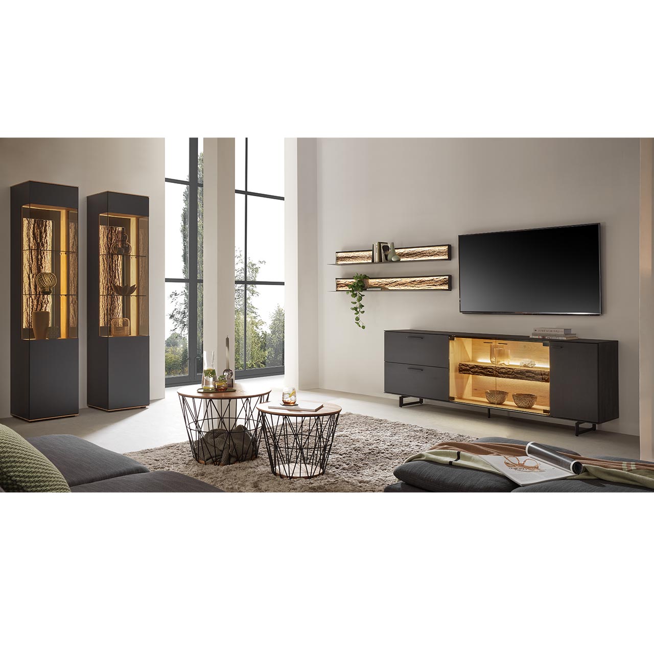Modernes Wohnzimmer mit Wohnwand bestehend aus einem Lowboard aus massiver Kerneiche Natur, anthrazit gebeizt mit beleuchtetem Glasausschnitt auf Metallkufen und zwei Wandregale aus Metall mit sichtbarer Baumrinde. Links zwei Standvitrinen.