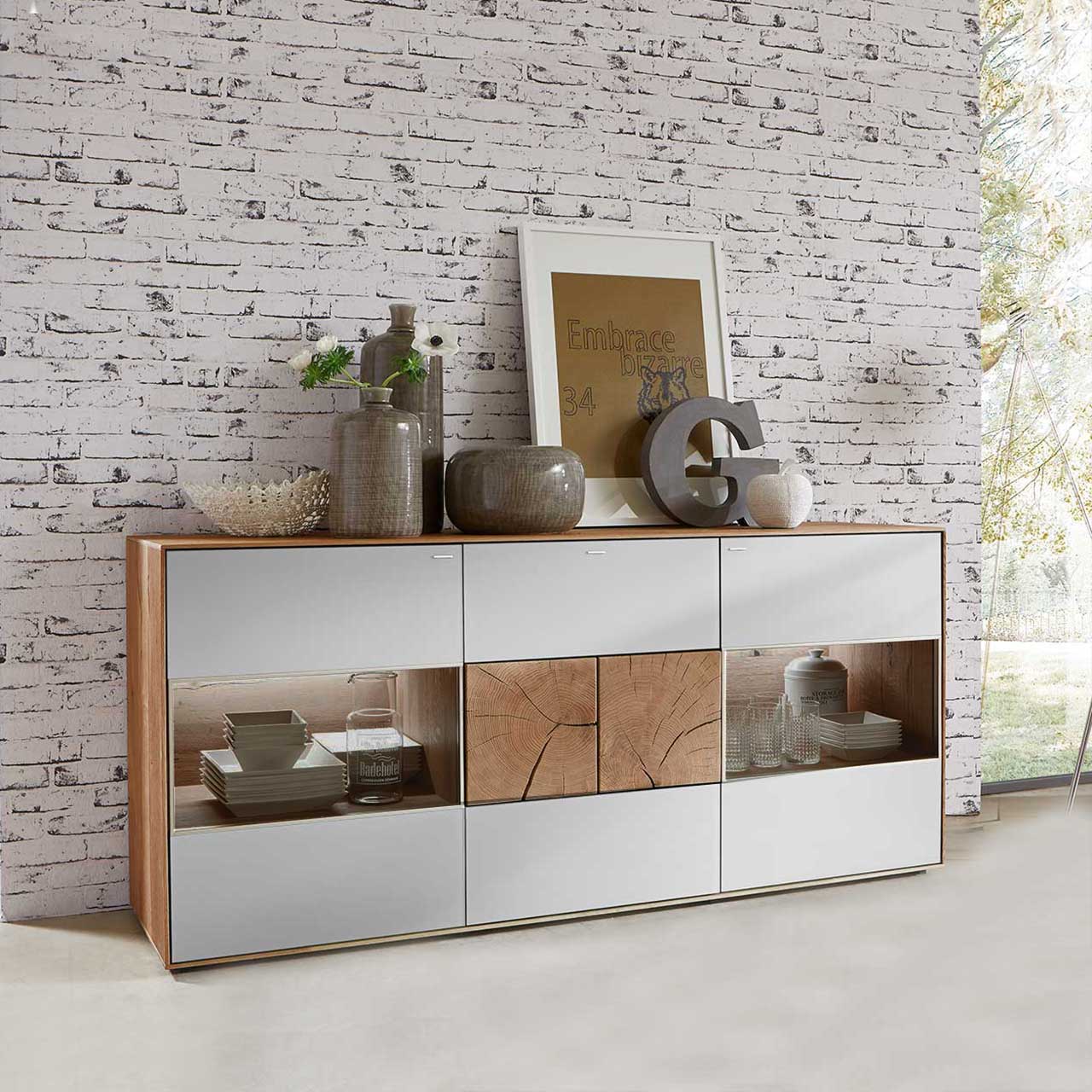 Milieubild mit modernem Sideboard aus massiver Kerneiche mit Mattglas weiß Fronten. Mit beleuchteten Glasausschnitten rechts und links und zwei rechteckigen Hirnholzscheiben in der Mitte - vor hellgrauer Steinwand-Tapete.