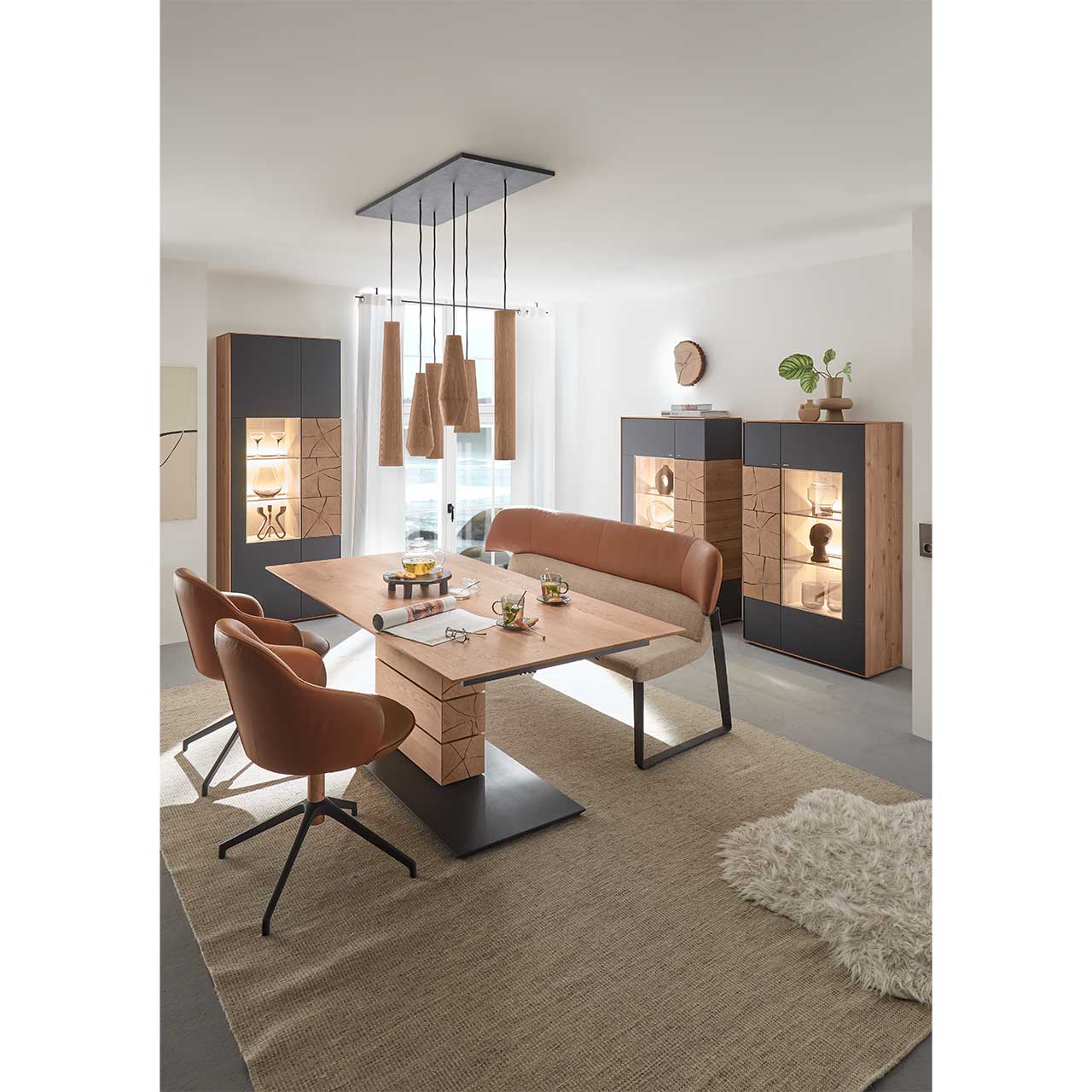 Modernes Esszimmer mit rechteckigem Massivholz Tisch mit Sitzbank und zwei Stühlen mit Lederbezug in cognac. Über dem Tisch hängt ein Pendelleuchte mit Holz-Lampenschirmen. Im Hintergrund stehen zwei passende Highboards und eine Standvitrine in Anthrazit.