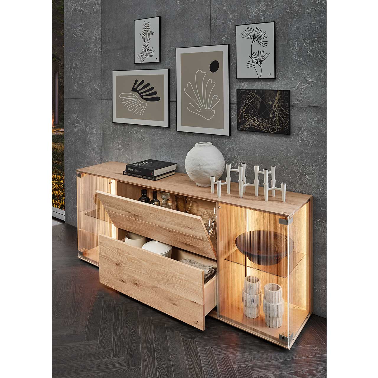 Milieubild eines Sideboards aus massiver Kerneiche Natur mit zwei beleuchteten Glastüren aus satiniertem Glas, geöffneter Klappe und offenem Schubkasten – vor grauer Wand mit Wandbildern.