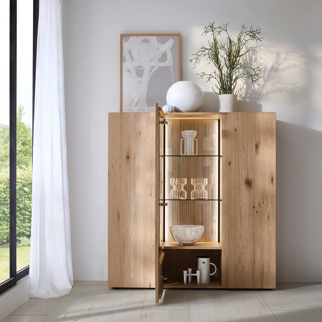 Modernes Highboard aus massiver Kerneiche Natur mit zwei Holztüren rechts und links und mittig mit offener Glas-/Holztür mit edel geschroppter Rückwand, zwei Glaseinlegeböden sowie einem Nischenfach unten - an grauer Wand stehend.