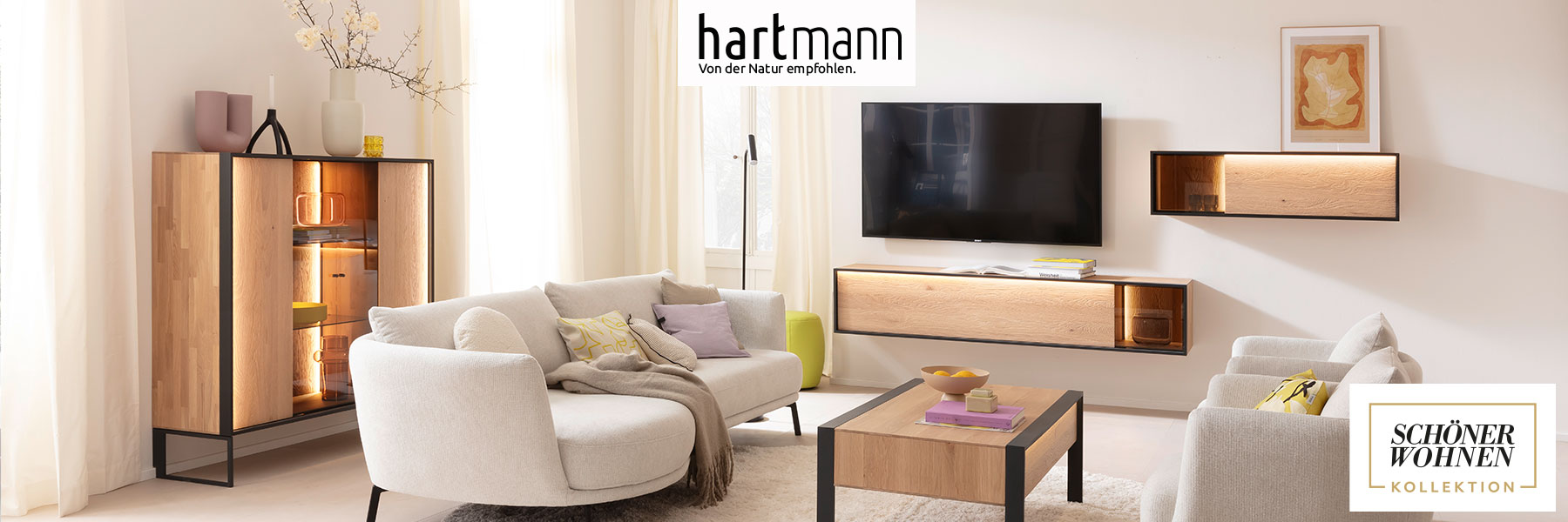 Hartmann Möbel - Möbelserie YOLA Schöner Wohnen Kollektion - Kerneiche Natur Hartmann YOLA - Wohnzimmermöbel aus massiver Kerneiche Natur sandgestrahlt kombiniert mit Metall anthrazit. Ambientebild zeigt ein beleuchtetes Hängelowboard und Hängevitrine, davor Couchtisch, links passendes Highboard mit Glasfront.