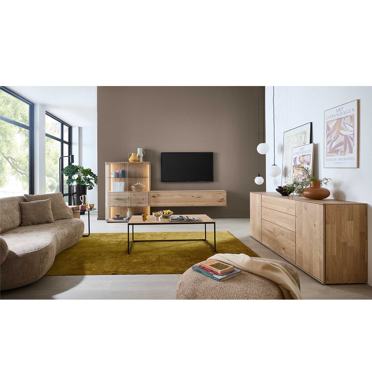 Modernes Wohnzimmer mit zweiteiliger Wohnwand aus Kerneiche Natur vor mokkafarbener Wand: Highboard mit großen Glastüren und Hängelowboard. Rechts das passende massive Sideboard. Im Vordergrund ein eckiger Couchtisch mit Metallgestell.