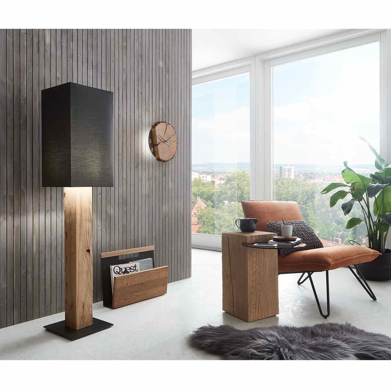 Wohnzimmer mit Stehlampe mit Stofflampenschirm und Holzfuß, Holz-Zeitungsständer und passende Holzwanduhr - vor dunkelgrauer Wand. Davor kleiner Beistelltisch aus Massivholz mit Glasablage.