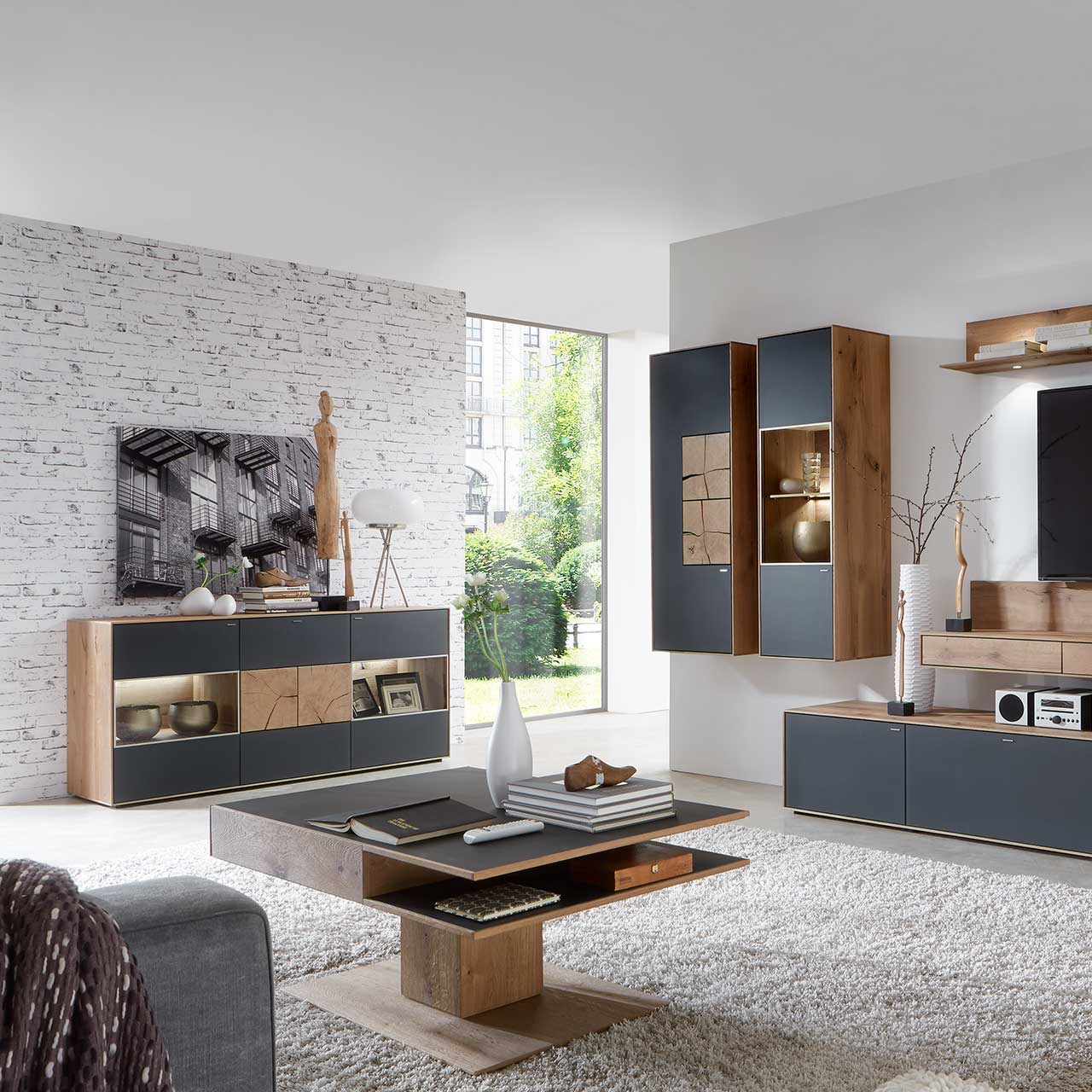 Wohnzimmer mit modernen Massivholzmöbel aus Kerneiche Umato mit Mattglas anthrazit Fronten: Hängeschrank mit Hirnholzscheiben, beleuchtete Hängevitrine, links modernes Sideboard mit Glasfront und Hirnholzscheiben, davor passender Couchtisch.