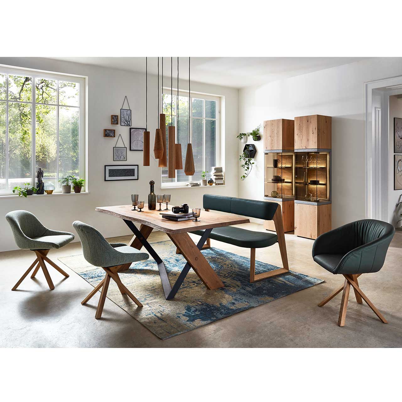 Modernes Esszimmer mit massivem Holztisch, drei Stühlen und einer Sitzbank mit dunkelgrünem Bezug und Holzgestell. Darüber eine moderne Pendelleuchte mit sechs unterschiedlichen geometrischen Holzkörpern. Im Hintergrund zwei Standvitrinen.