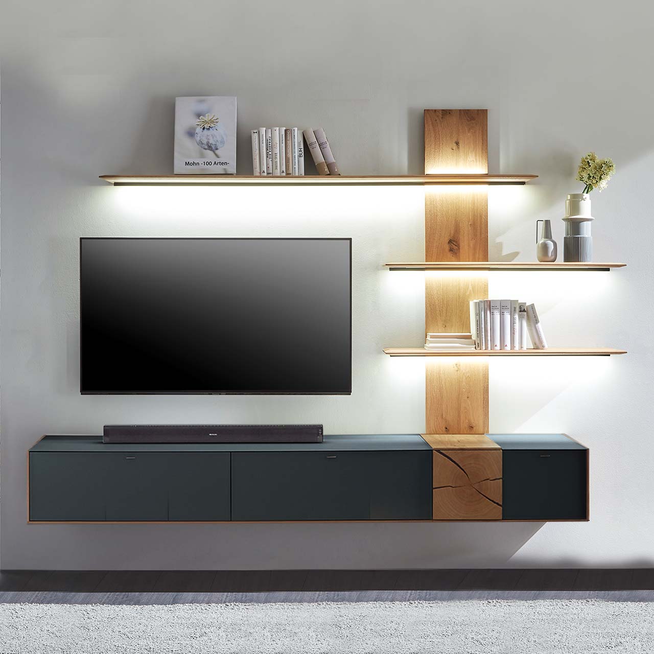 Moderne zweiteilige Massivholz Wohnkombination aus Kerneiche Umato mit Mattglas anthrazit Fronten und Hirnholzscheiben, mit einem langen Hängelowboard und einem Wandpaneel mit drei beleuchteten Regalböden. Dazwischen hängender Fernseher.