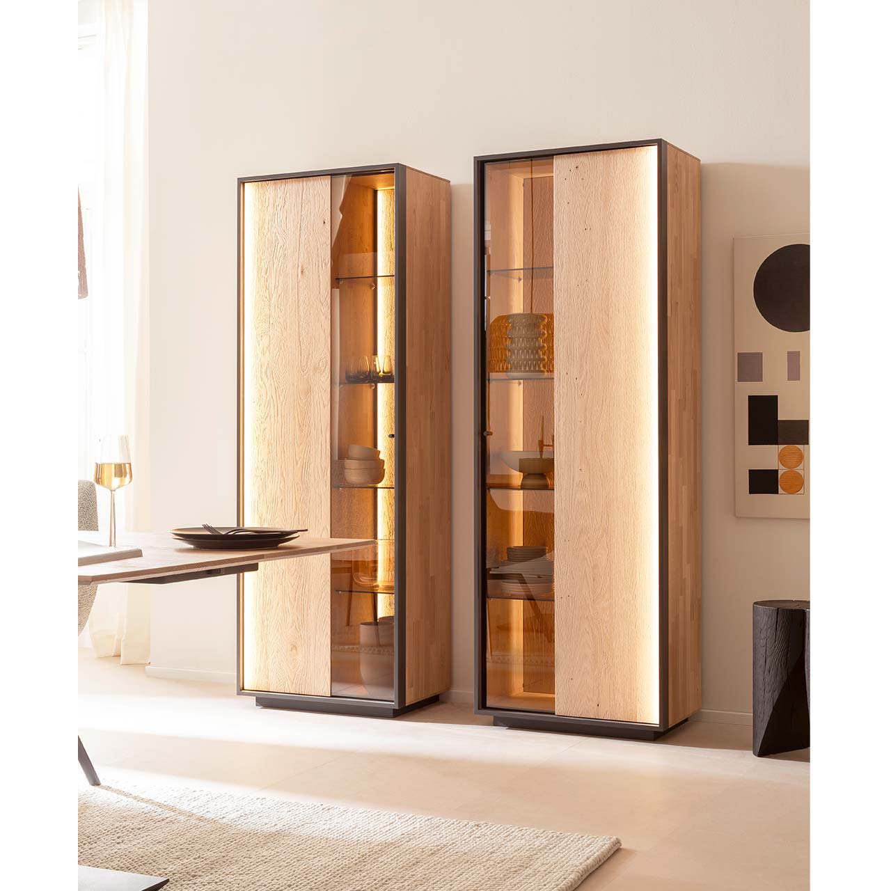 Zwei Standvitrinen mit Sockel im modernen Design aus sandgestrahlter Kerneiche Natur jeweils mit beleuchteter Holz-/Glastür, eingefasst in einem Metallrahmen in Anthrazit - vor einer hellbeigen Wand stehend.