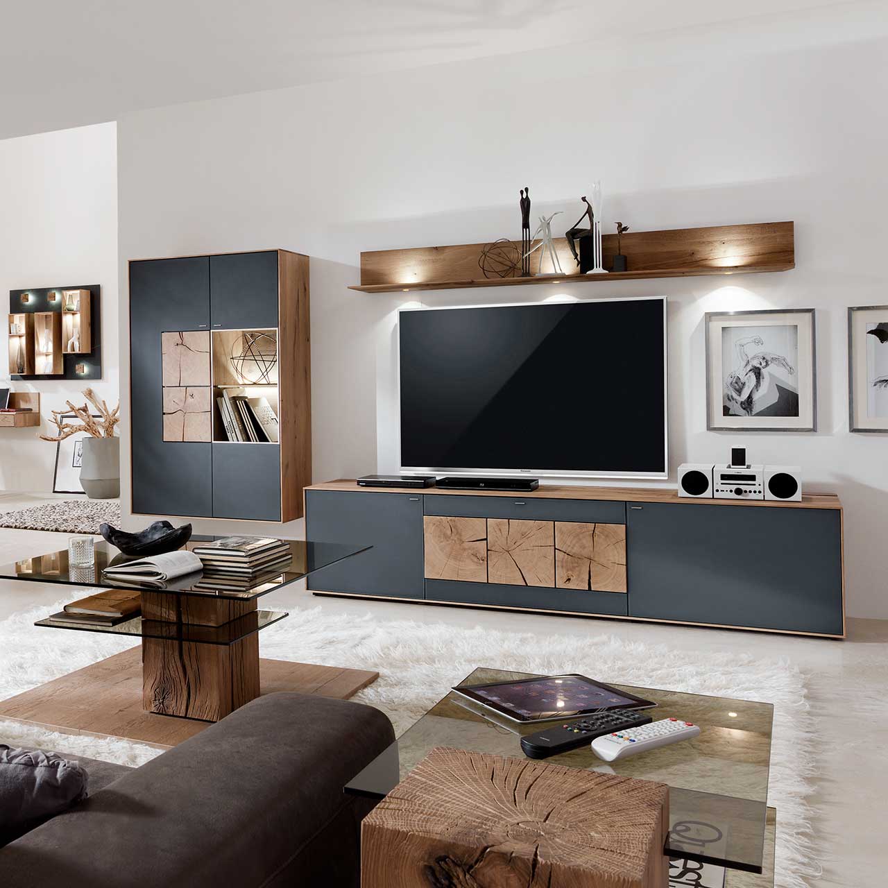 Wohnzimmer mit TV und moderner Wohnwand aus massiver Kerneiche Umato mit Mattglas anthrazit Fronten: Langes Lowboard mit dekorativen Hirnholzscheiben, Hängeregal aus Kerneiche, Hängevitrine mit beleuchtetem Glasausschnitt und Hirnholz.