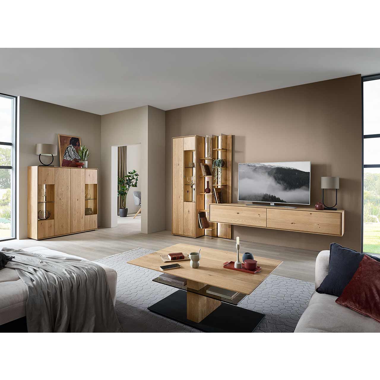 Modernes Wohnzimmer mit dreiteiliger Massivholzwohnwnad bestehend aus Standvitrine, hohem Wandregal mit Holzböden, beleuchteten Glasseiten und geschroppter Rückwand und einem darin integriertem Hängelowboard. Davor Couchtisch, links passendes Highboard.