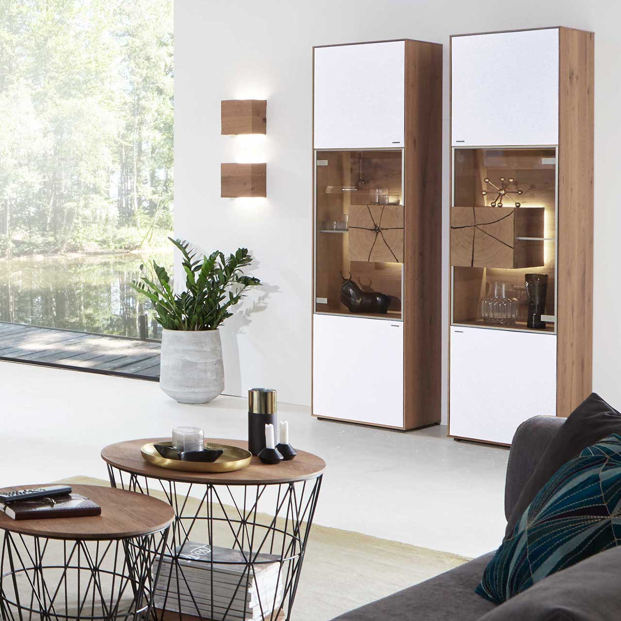 Wohnzimmerwand mit zwei modernen Standvitrinen aus massiver Kerneiche Umato mit Mattglas weiß Front, mittig mit beleuchtetem Glasausschnitt, Glasboden und Hirnholzblock. Links zwei Wandleuchten aus hellem Holz, im Vordergrund zwei runde Couchtische.