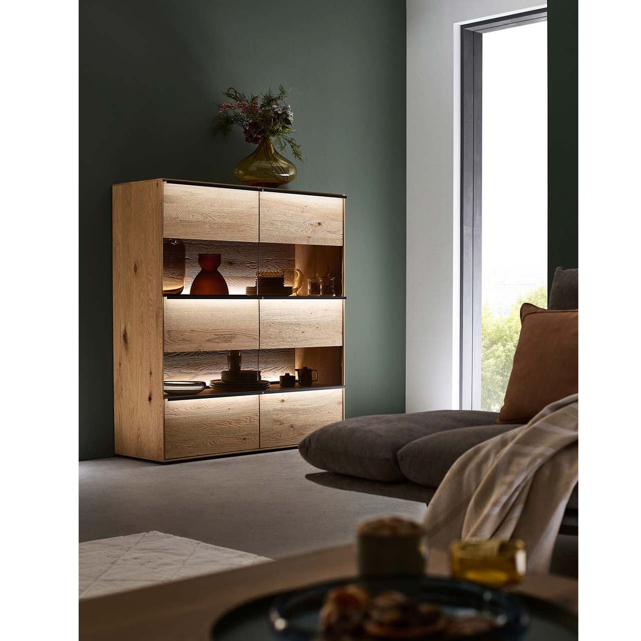 hartmann_ANEA_8530-7131_highboard_milieu1 Massivholz-Highboard mit 2 Türen inkl. Glasausschnitt mit optionaler Griffbeleuchtung vor einer dunkelgrünen Wand
