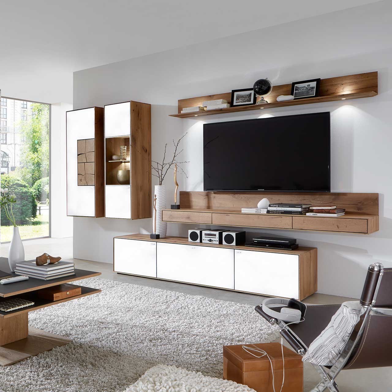 Wohnzimmer mit TV und moderner Wohnwand aus massiver Kerneiche Umato mit Mattglas weiß Fronten: Beleuchtetes Wandregal, langes Lowboard, beleuchtete Hängevitrine und Hängeschrank mit dekorativen Hirnholzscheiben.