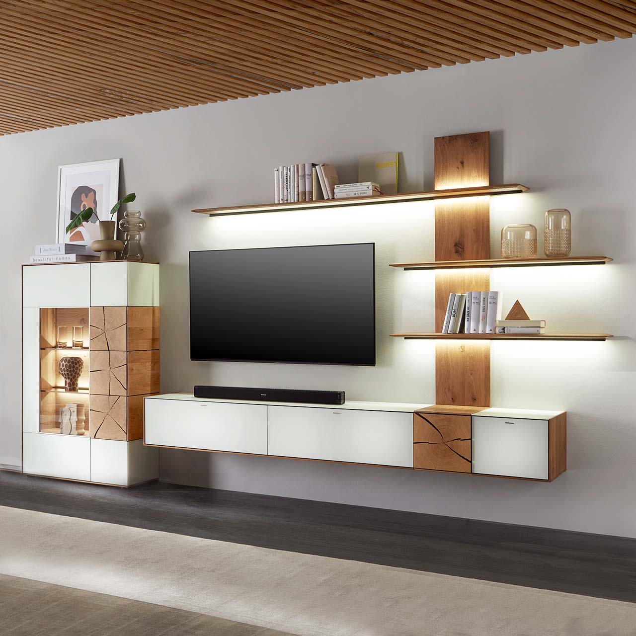 Massivholz Wohnwand aus Kerneiche Umato mit Mattglas weiß Fronten. Unten ein Hängelowboard mit dekorativer Holzapplikation, seitlich ein Wandpaneel mit beleuchteten Regalböden, links passende Standvitrine, dazwischen hängender Fernseher.