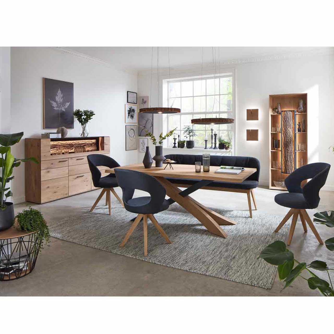 Modernes Esszimmer mit massivem, rechteckigem Holztisch, zwei Stühlen auf Holzgestell und passender Sitzbank in schwarzem Leder. Darüber hängend zwei Baumscheiben-Leuchten. Im Hintergrund die passenden Esszimmermöbel.