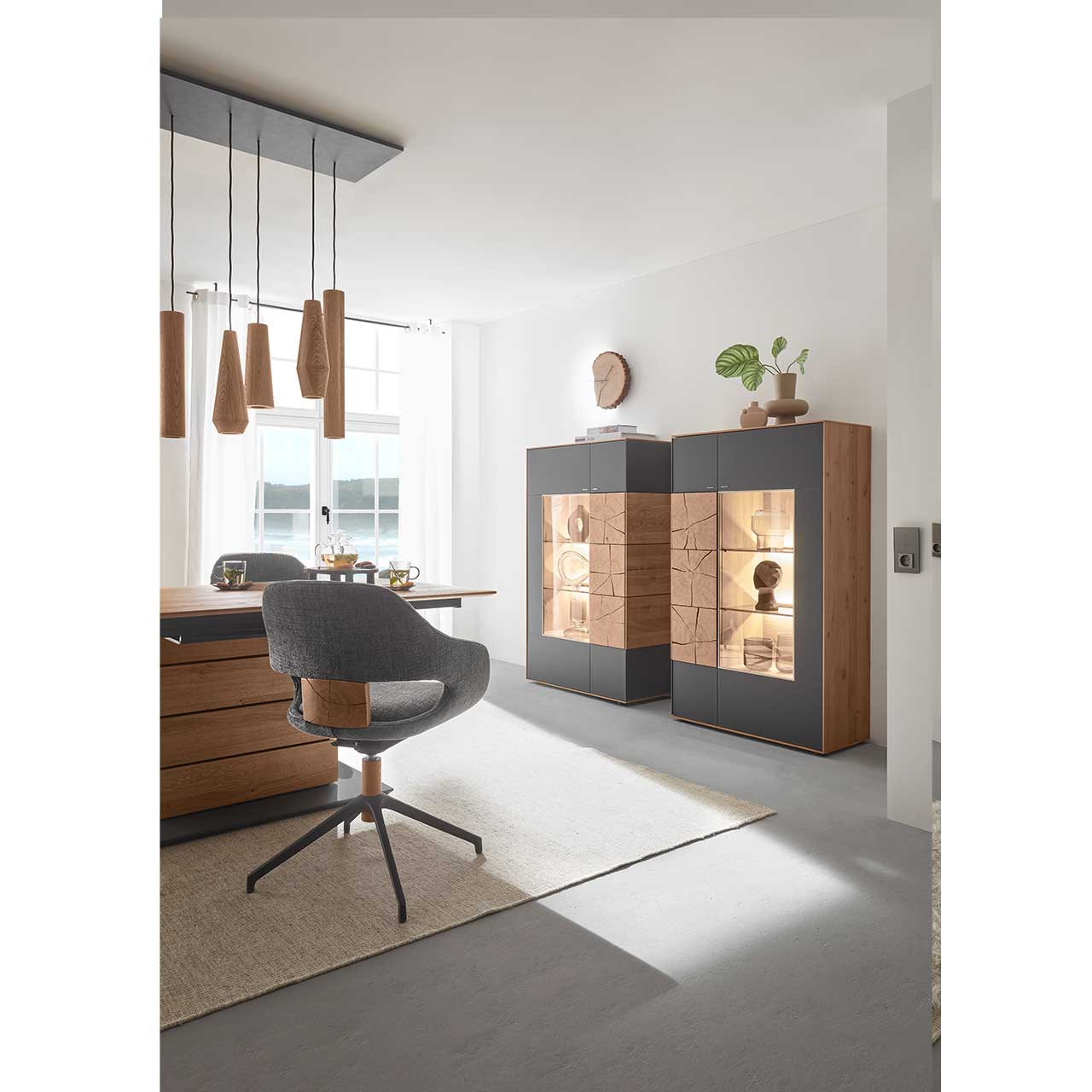 Modernes Esszimmer mit Massivholz Tisch und Stuhl in dunklem Stoff und Metallgestell. Über dem Tisch hängt eine Pendelleuchte mit Holz-Lampenschirmen. Seitlich an der Wand stehen zwei Highboards in Mattglas Anthrazit mit Glastüren und Hirnholzscheiben.