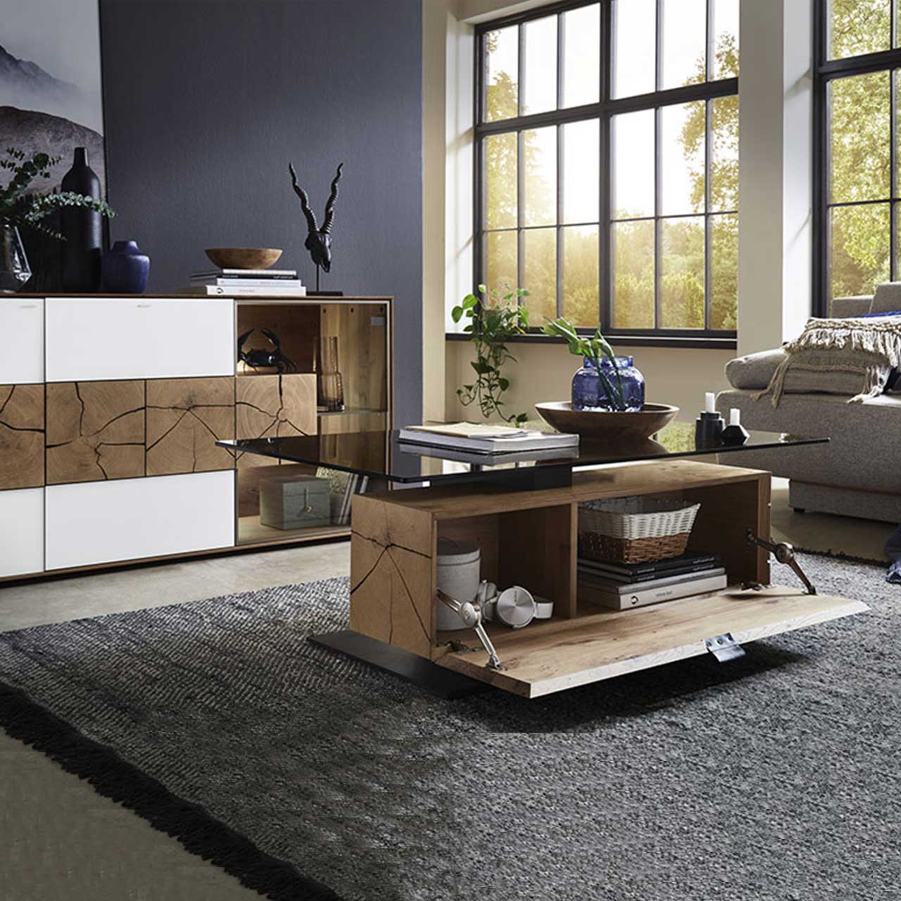 Bildausschnitt eines Wohnzimmers mit modernem Lowboard aus massiver Kerneiche Umato mit weißfarbenen Mattglasfronten, drei rechteckigen Hirnholzscheiben und Glastür. Davor passender Couchtisch mit Glasplatte und geöffnetem Stauraumfach.