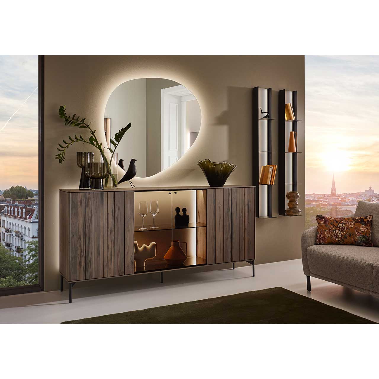 Wohnwand mit modernem Sideboard aus dunkelbrauner Altholzbuche Uyaa mit edler Holzmaserung, mittig mit Glasausschnitt und Glasboden vor beleuchteter Rückwand in Lack Sand - auf Metallfüßen. Darüber ein asymmetrischer Spiegel, rechts zwei Metallregale.