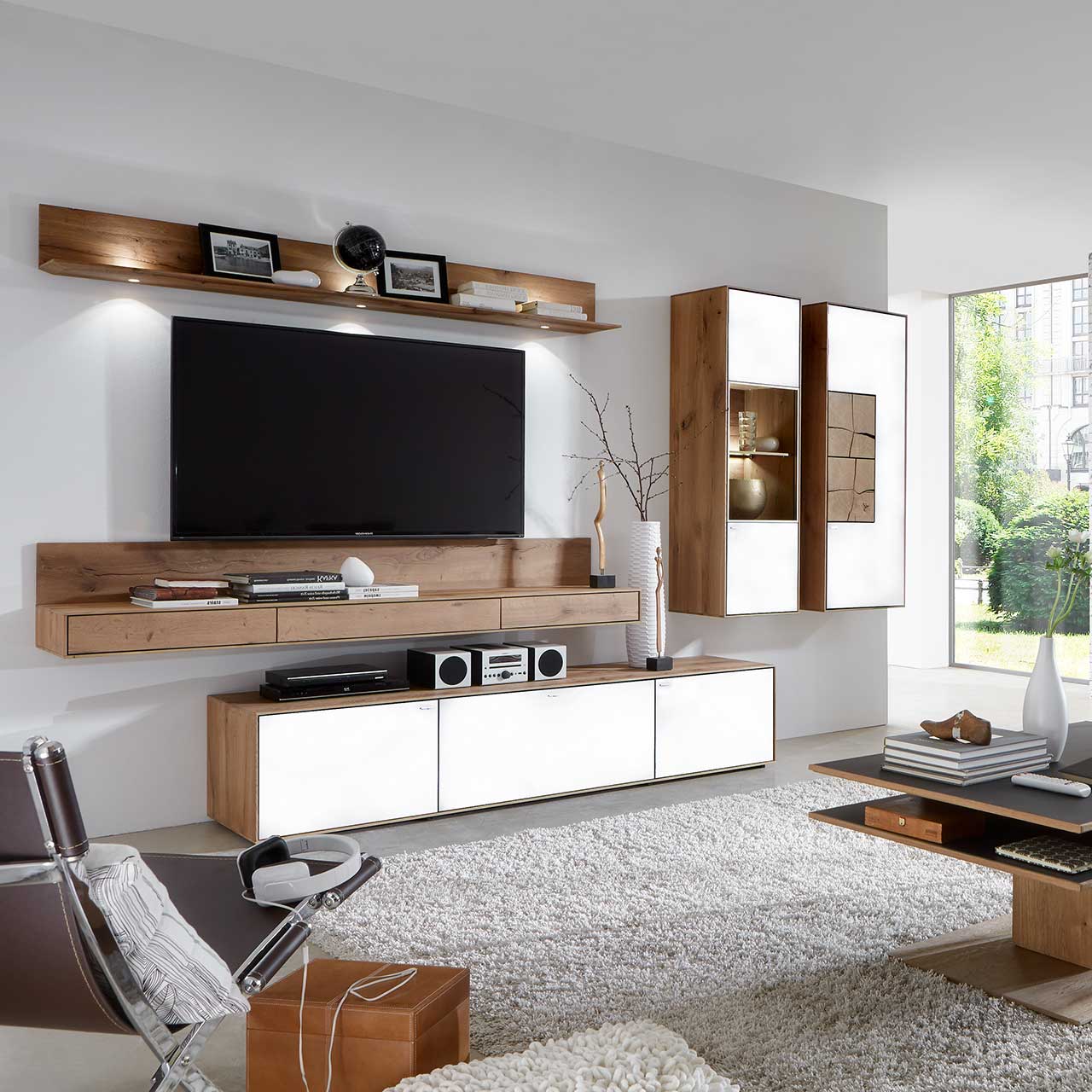 Milieubild Hartmann CAYA - Hängeelement 7170-7048W re. - Türanschlag rechts Wohnzimmer mit TV und moderner Wohnwand aus massiver Kerneiche Umato mit Mattglas weiß Fronten: Beleuchtetes Wandregal, langes Lowboard, beleuchtete Hängevitrine und Hängeschrank mit dekorativen Hirnholzscheiben.