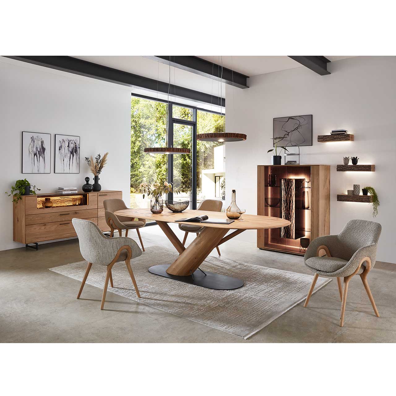 Modernes Esszimmer mit massivem, ovalem Holztisch und passenden Stühlen mit grauem Bezug und Holzgestell. Darüber hängend zwei Baumscheiben-Leuchten. Im Hintergrund die passenden Esszimmermöbel.