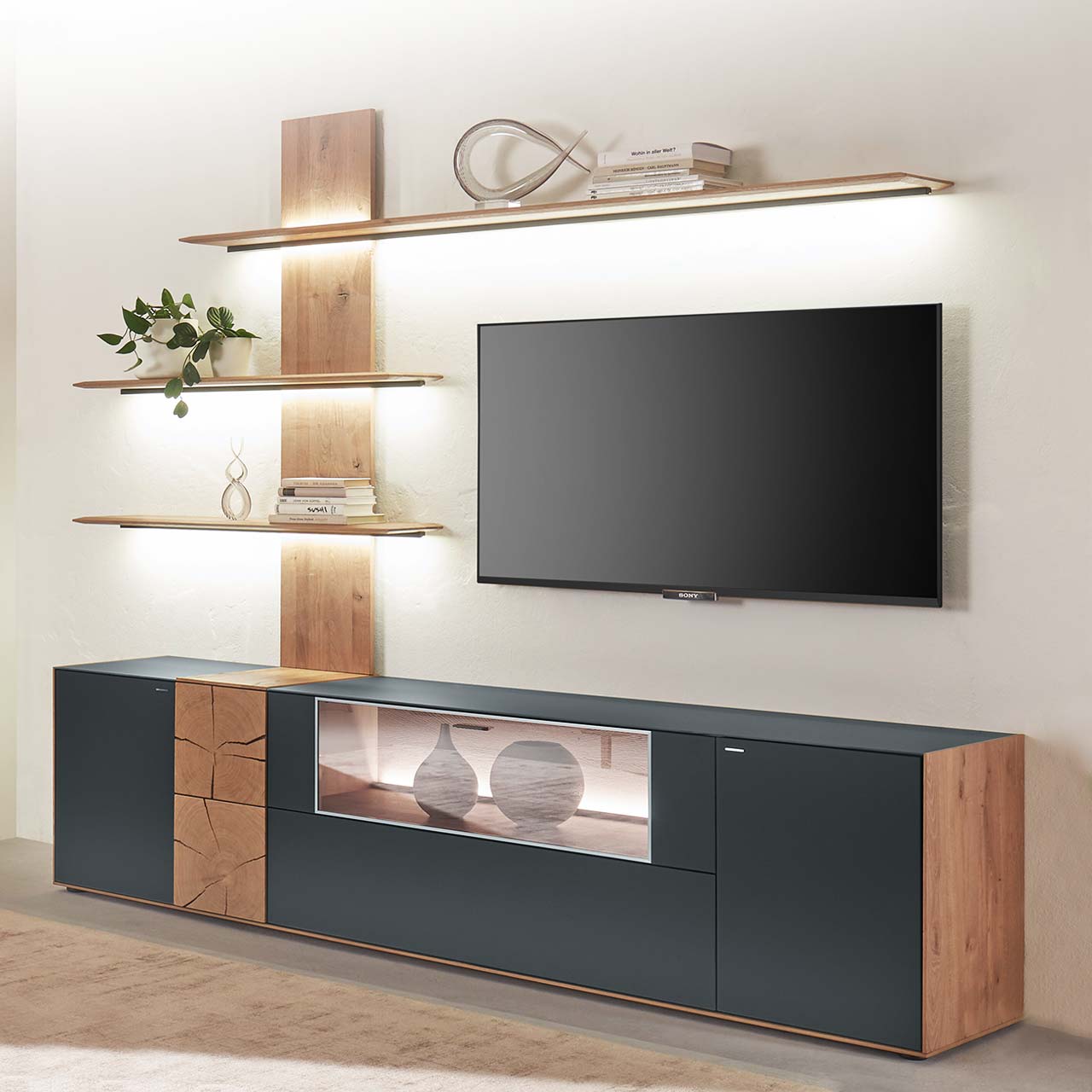 Moderne zweiteilige Massivholz Wohnkombination aus Kerneiche Umato mit Mattglas anthrazit Fronten und Hirnholzscheiben, mit einem langen Lowboard mit Glasausschnitt und einem Wandpaneel mit drei beleuchteten Regalböden. Dazwischen hängender Fernseher.