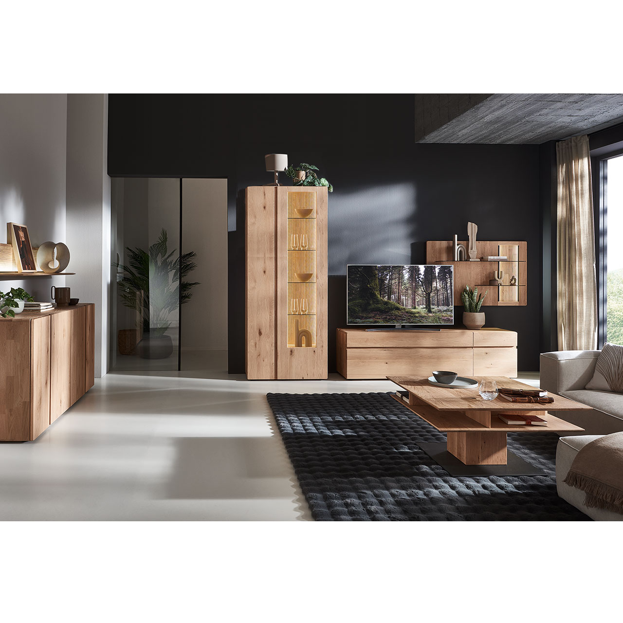 Modernes Wohnzimmer mit dreiteiliger Wohnwand aus massiver Kerneiche Natur: Lowboard, rechts Wandpaneel mit beleuchteten Glasböden sowie geschroppter Rückwand, links Standvitrine mit Holz-/Glastüren. Davor passender Holzcouchtisch, links Sideboard.