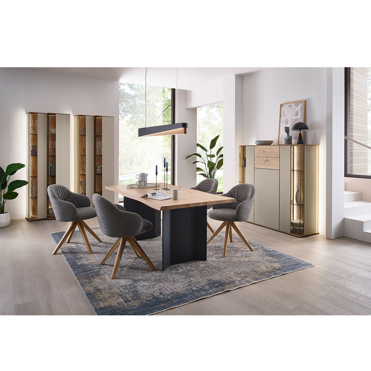 Hartmann Esszimmermöbel SVEA 8660 - aus massiver Kerneiche Natur, Lack Sand, Riffholz - Milieubild Esszimmermöbel aus massivem Holz mit Lackfronten in Sand, Parsolglas Bronze und Riffholz: Rechteckiger Holztisch mit Gestellwangen, vier Stühle mit dunkelgrauem Stoffbezug auf Holzgestell. Passendes Highboard und zwei Standvitrinen in Holz/Lack.